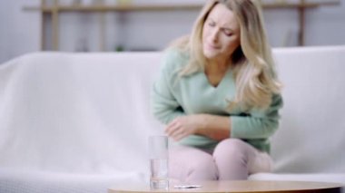 Bir bardak suyun seçici odağı ve arka planda karın ağrısı olan bir kadın. 