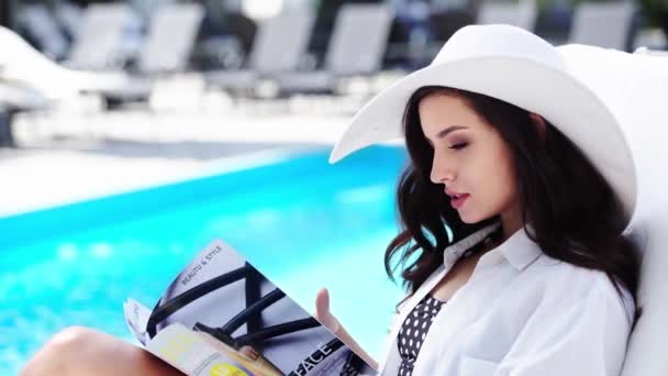 在游泳池附近的日光浴室里 戴着草帽的漂亮姑娘正在看杂志 — 图库视频影像