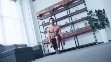 Gömleksiz bir sporcunun dambıllarla evde çömelmesi.