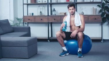 Yorgun, üstsüz sporcu spor topu üzerinde oturup su içiyor.
