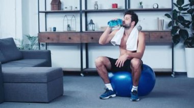 Spor topu üzerinde oturan yorgun sporcu ve spor şişesinden su içiyor.