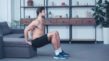 Kaslı, üstsüz sporcu evde kanepenin yanında çömeliyor.