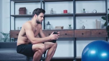 Üstsüz, yorgun sporcu kanepede oturuyor ve spor şişesinden içiyor.