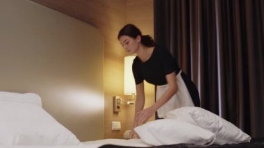 Genç, güzel üniformalı ve önlüklü hizmetçi otel odasında yatak yapıyor.