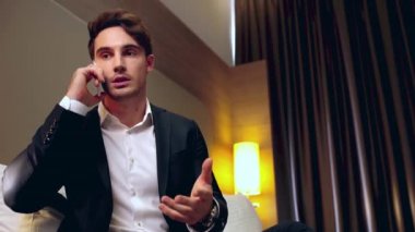Kendine güvenen, gülümseyen iş adamı otel odasında akıllı telefondan konuşuyor.