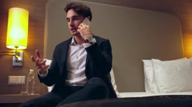 Gülümseyen iş adamı yatakta oturuyor ve otel odasında akıllı telefondan konuşuyor.