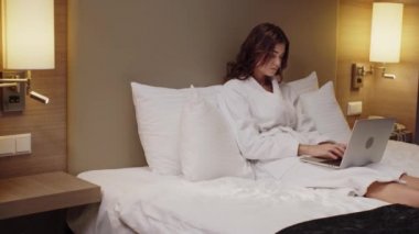 Bornozlu genç bir kadın otel odasında dizüstü bilgisayarında daktilo kullanıyor.