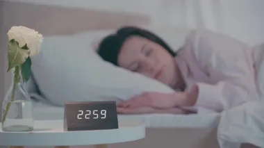Saatin seçici odak noktası ve gece yatakta uyuyan kadının yanındaki masada gül.