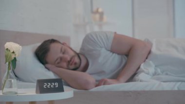 Yatakta uyuyan adamın yanındaki masada saatin ve çiçeğin seçici odağı