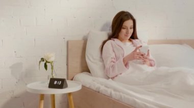 Sabahları pijamalı gülümseyen kız yatakta akıllı telefon kullanıyor. 
