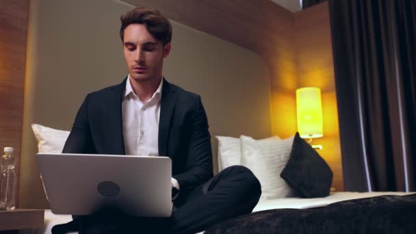 Νεαρός Σοβαρός Επιχειρηματίας Που Κάθεται Στο Κρεβάτι Και Χρησιμοποιεί Φορητό — Αρχείο Βίντεο