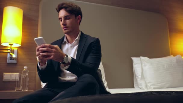 Gülümseyen Adamı Yatakta Oturmuş Otel Odasında Akıllı Telefondan Sohbet Ediyor — Stok video
