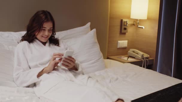 幸せな女の子ですバスローブの女の子はベッドで休んでいる間にスマートフォンでチャット — ストック動画