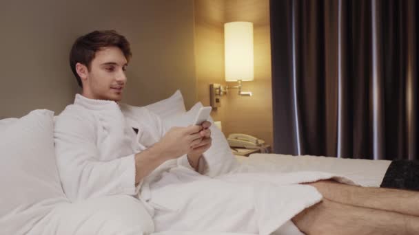 Jeune Homme Concentré Souriant Tout Reposant Lit Bavardant Sur Smartphone — Video