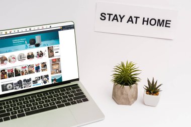 KYIV, UKRAINE - 8 Nisan 2020: bitkilerin ve kağıdın yanında Amazon web sitesi olan dizüstü bilgisayar beyaz harflerle evde kalıyor  