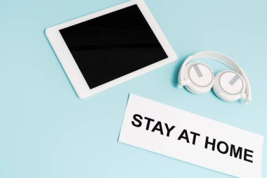 digital tablet with blank screen near headphones and paper with stay at home lettering on blue  clipart