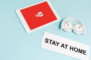 KYIV, UKRAINE - APRIL 8, 2020: digital tablet with youtube app near headphones and paper with stay at home lettering on blue  clipart
