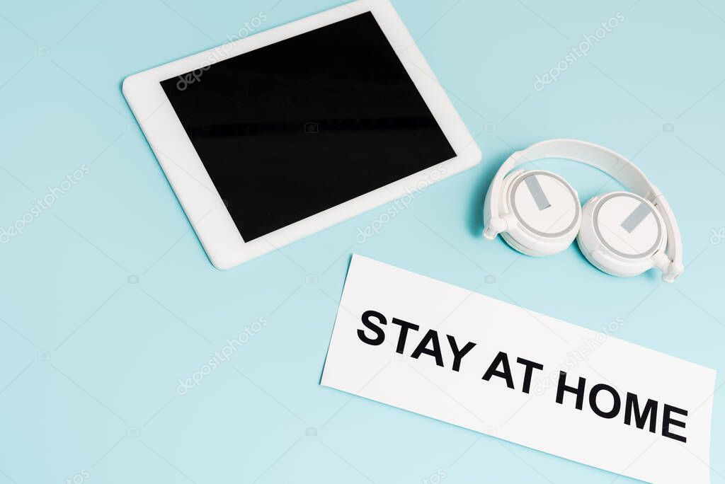 digital tablet with blank screen near headphones and paper with stay at home lettering on blue 