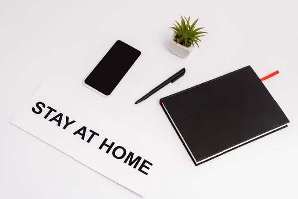 Smartphone con pantalla en blanco cerca de bolígrafo, cuaderno, planta y papel con letras de estancia en casa en blanco - foto de stock