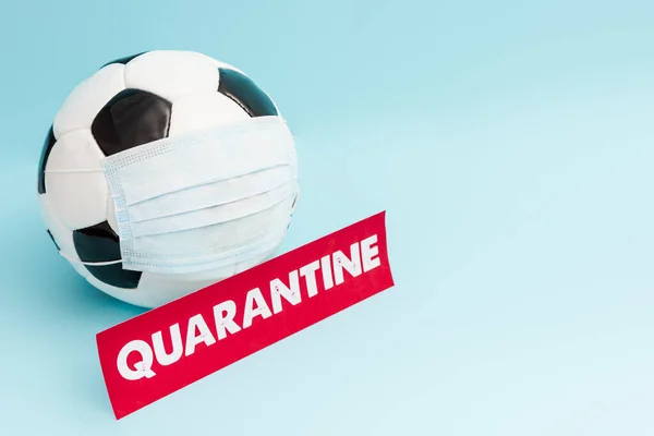 Ballon de football en masque médical près du papier avec lettrage de quarantaine sur bleu — Photo de stock