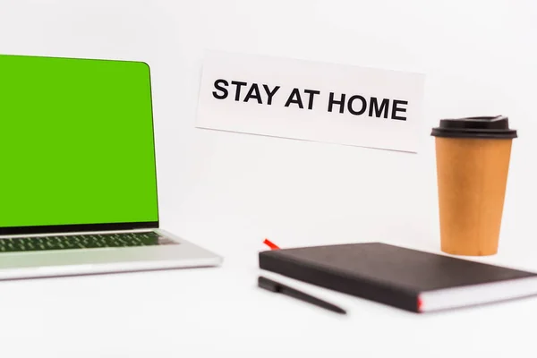 Enfoque selectivo de la computadora portátil con pantalla verde cerca de la taza de papel, cuaderno, pluma y papel con la estancia en casa letras aisladas en blanco - foto de stock