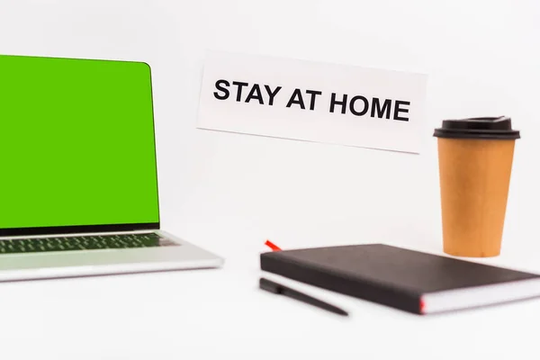 Messa a fuoco selettiva del computer portatile con schermo verde vicino a tazza di carta, notebook, penna e carta con soggiorno a casa lettering isolato su bianco — Foto stock