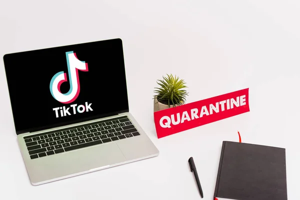 KYIV, UKRAINE - 8 AVRIL 2020 : ordinateur portable avec tik tok site web à l'écran près de la plante verte, stylo, ordinateur portable et papier avec lettrage de quarantaine sur blanc — Photo de stock