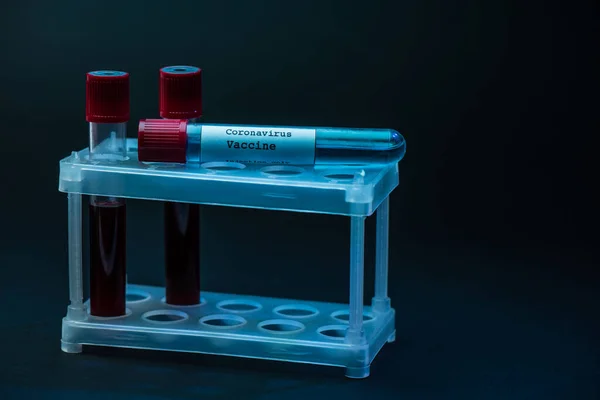 Vacuna contra el Coronavirus en el estante del tubo de ensayo con tubos de muestra con sangre sobre fondo oscuro - foto de stock