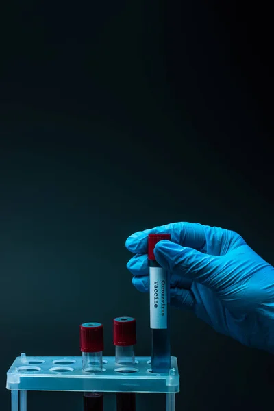 Vista recortada de la mano masculina con la vacuna contra el coronavirus y tubos de muestra con sangre en el estante del tubo de ensayo sobre fondo oscuro - foto de stock