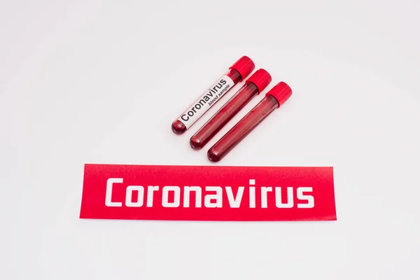 Vista de alto ângulo do cartão com letras coronavírus perto de tubos de ensaio com amostras de sangue em branco — Fotografia de Stock