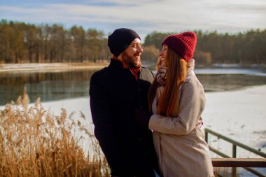 Couple in a winter forest and frozen lake. Young lovers in winter jackets laugh.