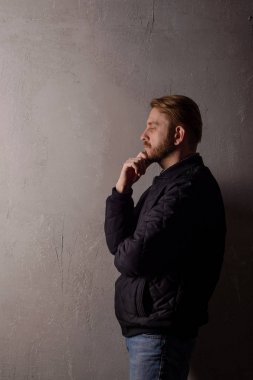 Pensive middle-aged bearded man in a black jacket and jeans stands near the wall looking away clipart