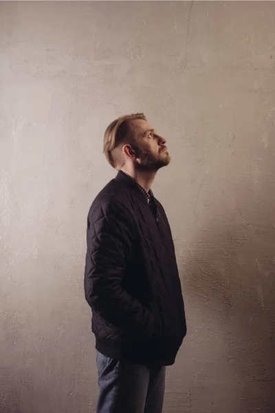 An attractive middle-aged bearded man in a black jacket and jeans stands behind the wall looking sideways to the top.