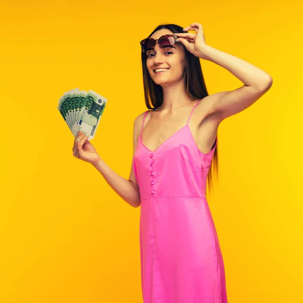 Slender Spanish girl in a pink dress and sunglasses holds a hundred euro bills on a yellow background