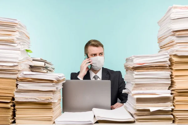 A male accountant or company manager works in an office in a pandemic in view of the accumulated paper work. A protective medical mask is on the face. On the desktop are large stacks of documents. - image