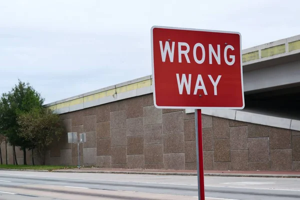 A Wrong Way sign on the side of a freeway frontage road in the city suburbs with an overpass in the background and empty streets.