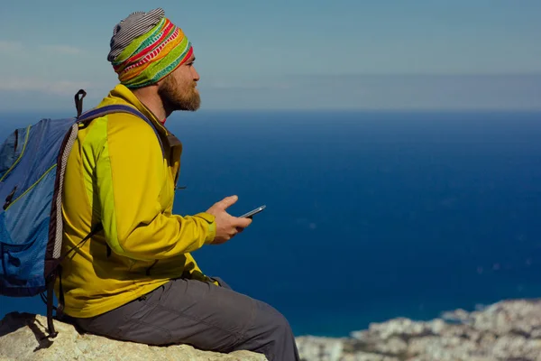 Ο άνθρωπος που κάθεται πάνω σε ένα βράχο πάνω από τη θάλασσα με ένα smartphone στο χέρι — Φωτογραφία Αρχείου