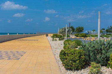 Marjan Island in Ras al Khaimah, United Arab Emirates boardwalk along the Arabian Gulf (Persian Gulf) on a blue sky day with green palm trees and plants clipart