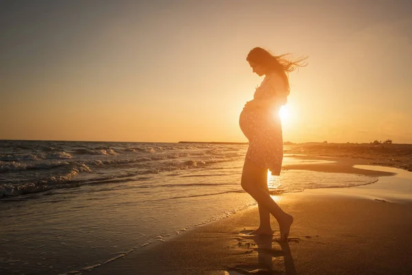 Silhueta de uma mulher grávida andando na praia ao pôr do sol — Fotografia de Stock