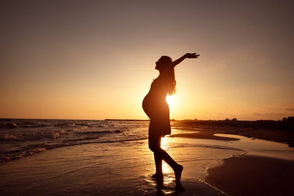 Silhueta de uma mulher grávida andando na praia ao pôr do sol — Fotografia de Stock