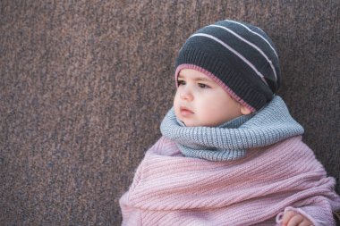 Bir sıcak kış şapka ve renkli bir eşarp kahverengi bir arka plan üzerinde giyen şirin bebek kız.