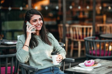 Genç bayan bir kafede oturuyor ve mutlu bir gülümsemeyle cep telefonuyla konuşuyor.