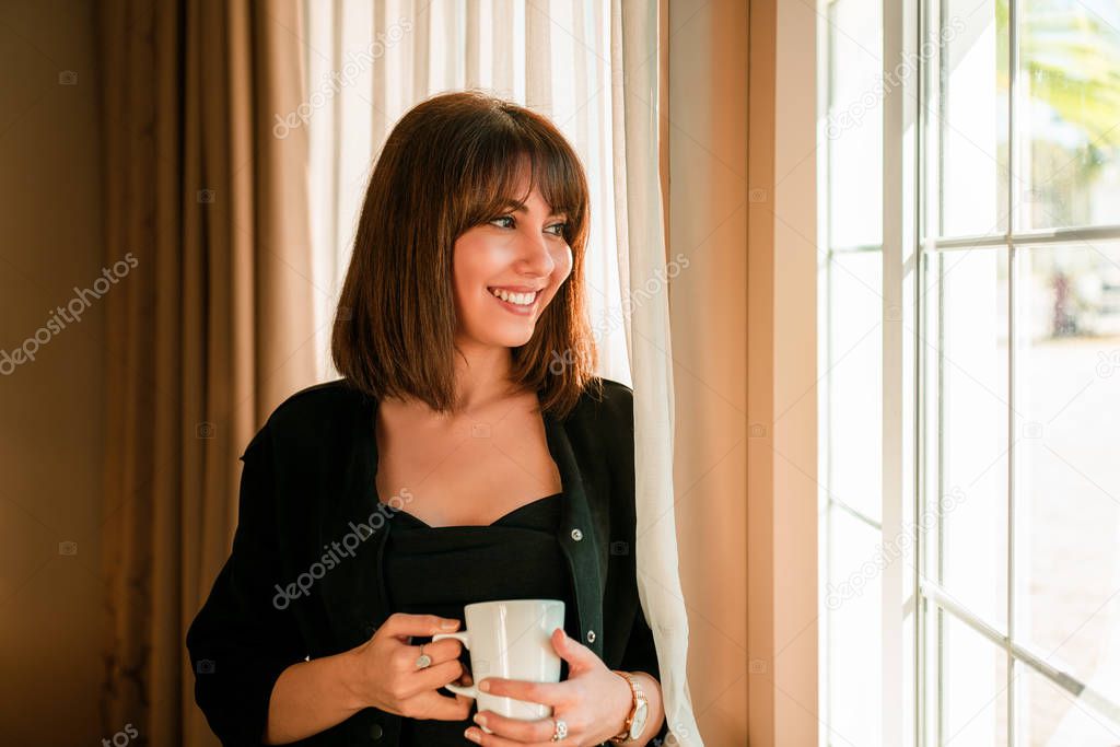 Young woman smiling and drinking some beverage and looking out from window