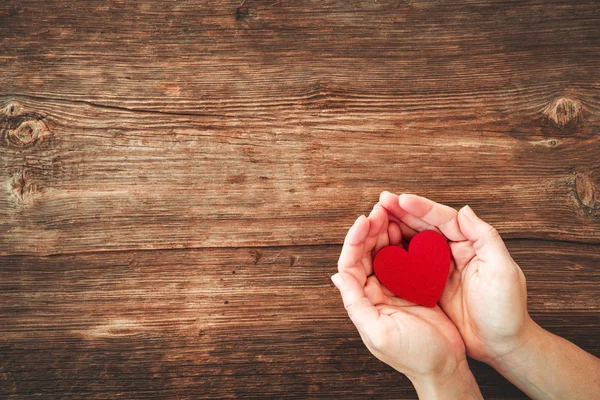 Vista superior de las manos de mujer sosteniendo el corazón rojo en la mesa de madera . — Foto de Stock