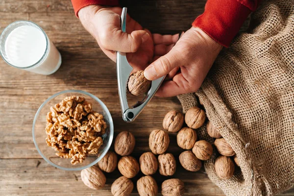 Close up top view of male hands cracking walnuts with nutcracker and a glass of milk on a wooden table. — Stock Photo, Image