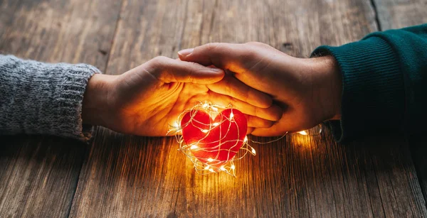 Dvě ruce držící červené srdce zakryté světly na dřevěném pozadí a bokeh světly. Valentýn a romantický koncept. — Stock fotografie