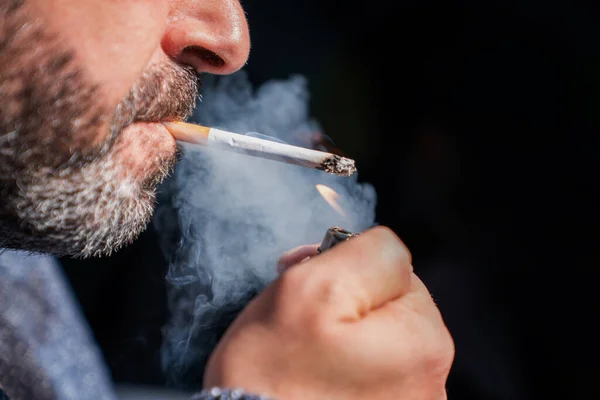 Nahaufnahme eines bärtigen Mannes, der ein Feuerzeug in der Hand hält und eine Zigarette auf schwarzem Hintergrund raucht. — Stockfoto