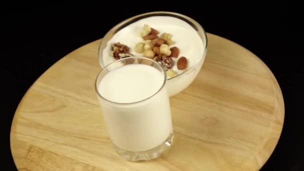 Yogurt con nueces y un vaso de leche rotan sobre una plataforma de madera sobre un fondo negro — Vídeos de Stock