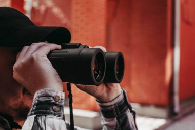 Sakallı bir adam şehirde dürbünle bakar.