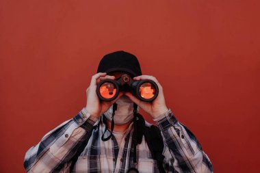 Gömlekli bir adam kameraya kırmızı arka plandaki dürbünle bakıyor.